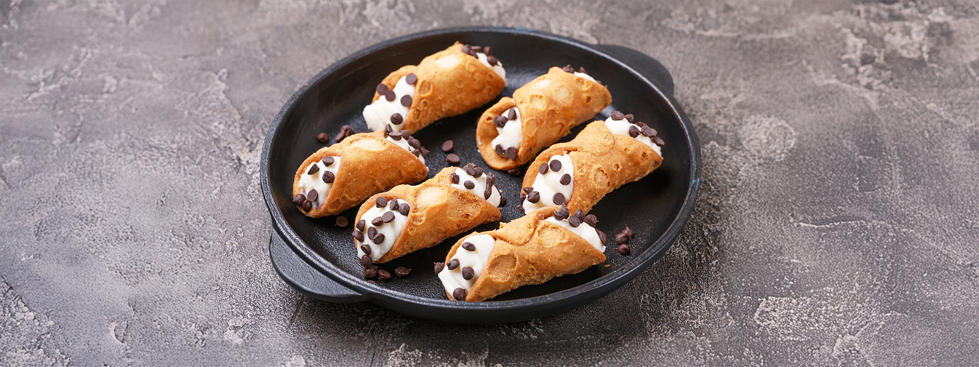 Piatto nero con 6 cannoli alla ricotta e gocce di cioccolato su piano grigio